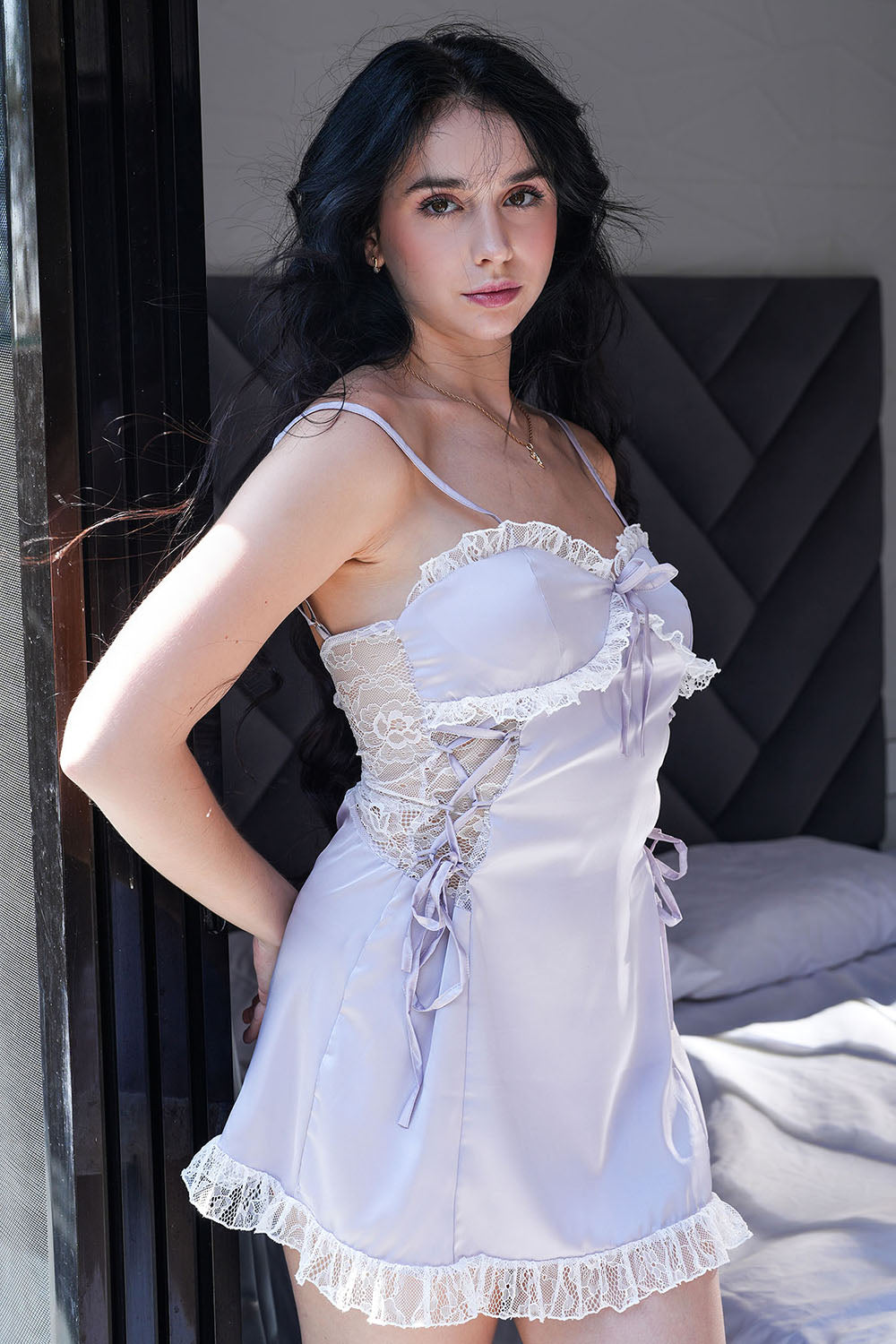 Woman standing indoors in lavender satin lace-trim babydoll dress, side-front view.