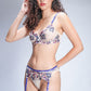 Model in sheer floral lingerie set with blue straps and garter belt, posing against grey background.