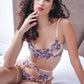 Woman sitting on bed in sheer floral lingerie with nude garter straps, striking a sensual pose.